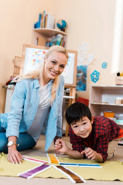 Selektiver Fokus von Lehrer und Kind, die in die Kamera lächeln, während sie in der Montessori-Schule auf dem Boden spielen — Stockfoto