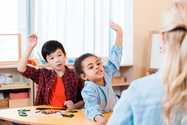 Enfoque selectivo de los niños con las manos levantadas y el maestro durante la lección en la clase montessori - foto de stock