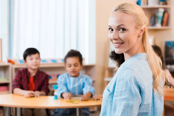 Focus selettivo dell'insegnante sorridente che guarda la macchina fotografica con i bambini sullo sfondo nella scuola montessori — Foto stock