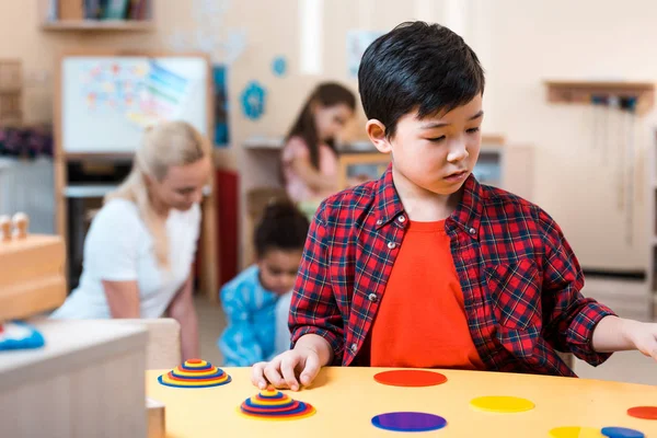 Focus selettivo del bambino asiatico pieghevole gioco e insegnante con i bambini in background nella scuola montessori — Foto stock