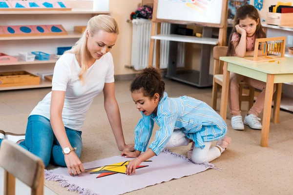 Focus selettivo dell'insegnante con bambino che gioca sul pavimento e bambino noioso alla scrivania nella scuola montessori — Foto stock