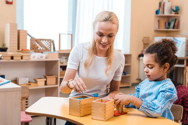 Profesor sonriente jugando juego de madera con el niño durante la lección en la escuela montessori - foto de stock