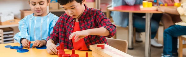 Tiro panorâmico de crianças jogando jogo de tabuleiro com professor e crianças em segundo plano na escola montessori — Fotografia de Stock