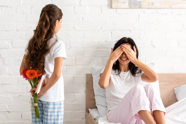 Vista posterior del niño sosteniendo las flores y la tarjeta de día de las madres mientras mamá se sienta en la cama y cubre los ojos con las manos - foto de stock