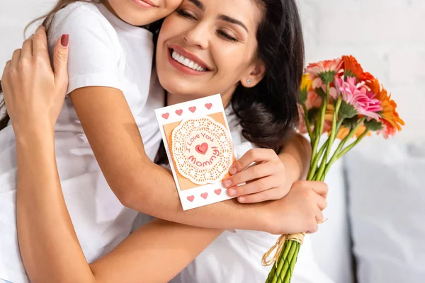Feliz mujer abrazando hija sosteniendo flores y madres día tarjeta con te amo mamá inscripción - foto de stock