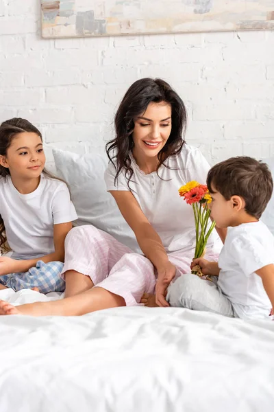 Menino adorável segurando flores enquanto sentado na cama perto de mãe feliz e irmã no dia das mães — Fotografia de Stock