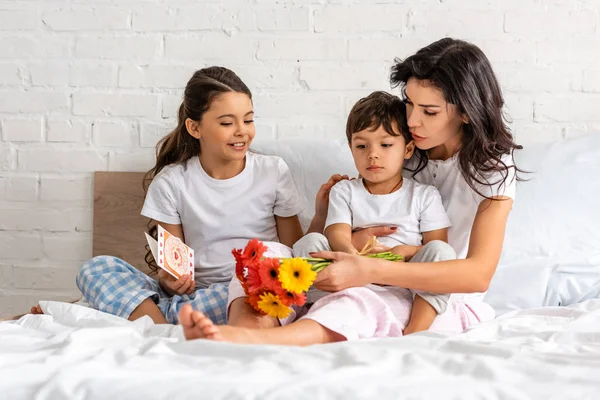 Mujer feliz sosteniendo flores y abrazando al hijo, mientras que la hija sosteniendo la tarjeta del día de las madres - foto de stock