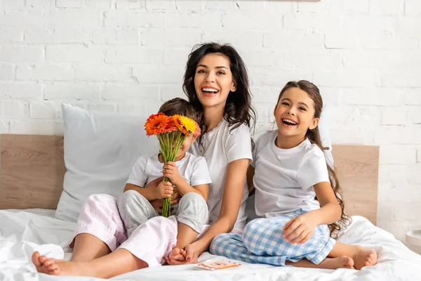 Cute boy holding flowers while sitting in bed near happy mother and sister — Stock Photo