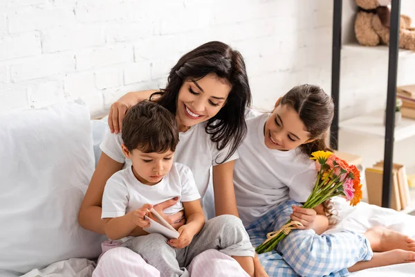 Bambino felice che tiene i fiori mentre si siede a letto con mamma e fratello il giorno delle madri — Foto stock