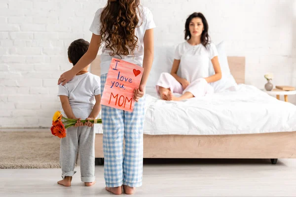 Back view of brother and sister holding flowers and i love you mom card, and surprised woman sitting in bed — Stock Photo