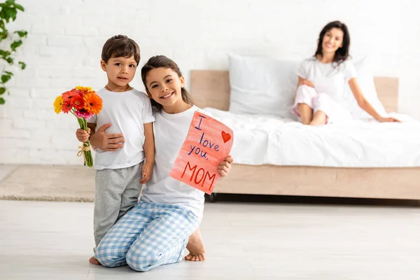 Enfoque selectivo de los niños sosteniendo te amo tarjeta de mamá cerca de la madre sentada en la cama - foto de stock