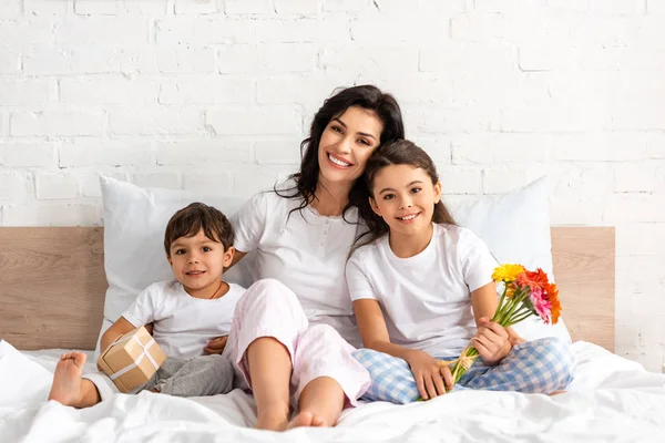 Femme heureuse avec des enfants souriant à la caméra tout en étant assis dans le lit avec des fleurs et boîte cadeau — Photo de stock