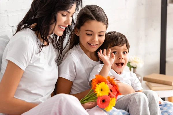 Lindo chico saludando mano cerca de madre y hermana sosteniendo flores en día de la madre - foto de stock