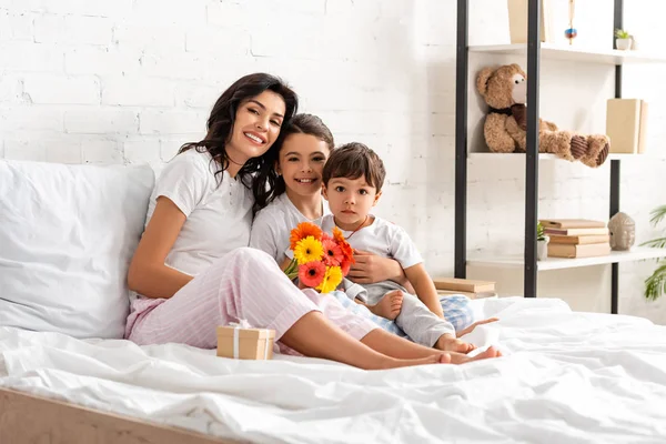 Felice madre abbracciare i bambini mentre seduto a letto con le madri giorno bouquet e confezione regalo — Foto stock