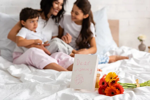 Selective focus of happy mother hugging adorable children while sitting in bed near bouquet and mothers day card with dear mommy lettering — Stock Photo