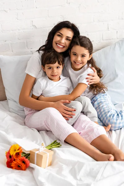 Madre feliz abrazando a los niños adorables mientras está sentada en la cama cerca del ramo y el regalo del día de las madres - foto de stock