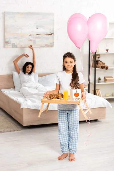 Cute daughter holding tray with breakfast, mothers day card with heart sign and mom lettering, while mother stretching in bed — Stock Photo