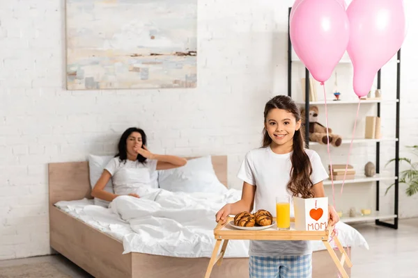 Vista recortada de la bandeja de sujeción del niño con desayuno, tarjeta de día de las madres con signo de corazón y letras de mamá, mientras que la madre estirándose en la cama - foto de stock