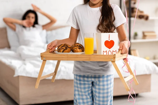 Cropped view of child holding tray with breakfast, mothers day card with heart sign and mom lettering, while mother stretching in bed — Stock Photo