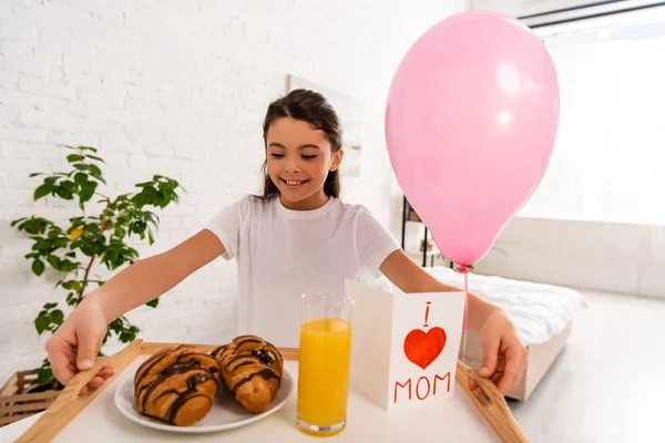 Happy child holding tray with croissants, orange juice and mothers day card with heart symbol and mom lettering — Stock Photo
