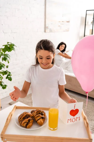 Criança feliz em pé perto da bandeja com café da manhã, cartão de dia das mães com sinal de coração e lettering mãe, enquanto a mãe sentada na cama — Fotografia de Stock