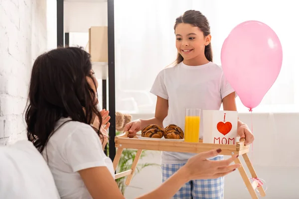 Sonriente hija sosteniendo bandeja con la tarjeta del día de las madres con el signo del corazón y las letras de mamá cerca de la madre sentada en la cama - foto de stock