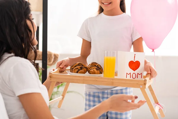 Vista recortada de la bandeja de retención de niños con desayuno, tarjeta de día de las madres con signo de corazón y letras de mamá cerca de la madre sentada en la cama - foto de stock