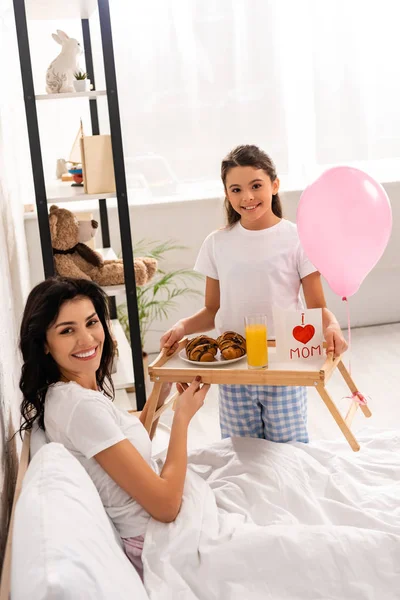 Cute child holding tray with breakfast, mothers day card with heart sign and mom lettering near happy mother sitting in bed — Stock Photo