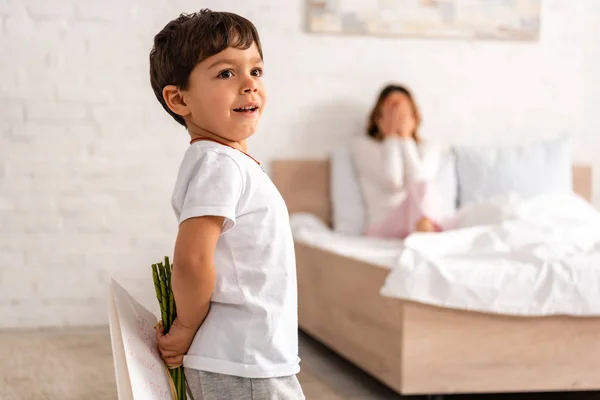 Foco seletivo de menino adorável segurando flores e mães cartão de dia, e mulher sentada na cama e cobrindo os olhos com as mãos — Fotografia de Stock