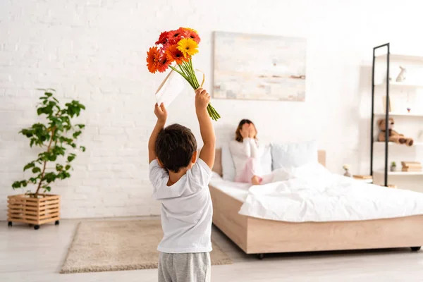 Back view of boy holding flowers and mothers day card in raised hands, and mother sitting in bed and covering eyes with hands — Stock Photo