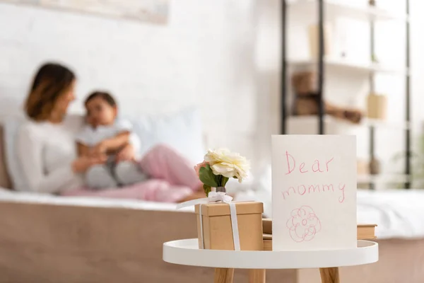 Selective focus of mothers day card with dear mommy inscription near gift box and roses on table, and happy woman sitting on bed with son — Stock Photo
