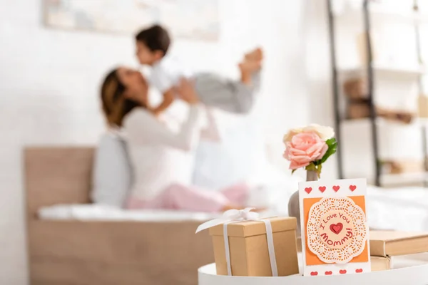 Foyer sélectif de la carte de la fête des mères avec je t'aime maman inscription et boîte cadeau sur la table, et femme heureuse s'amuser avec son fils au lit — Photo de stock