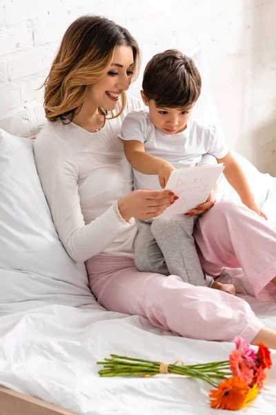 Happy mother holding adorable son and mothers day card while sitting in bed near flowers — Stock Photo