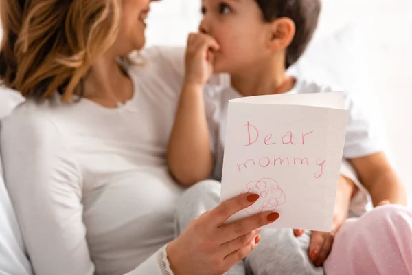 Foco seletivo da mulher feliz segurando adorável filho e mães cartão de dia com querida inscrição mamãe — Fotografia de Stock