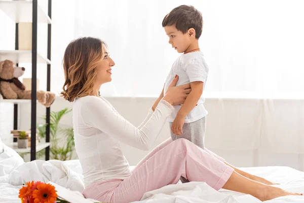 Vista lateral de la mujer feliz tocando adorable hijo mientras está sentado en la cama en el día de las madres - foto de stock