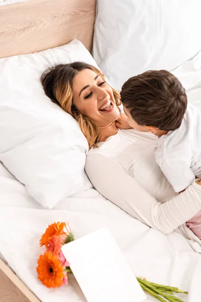 Cheerful woman having fun with son while lying in bed near flowers and mothers day card — Stock Photo