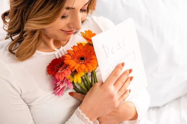 Glückliche Frau lächelt, während sie einen Strauß Gerberas und eine Muttertagskarte in der Hand hält — Stockfoto