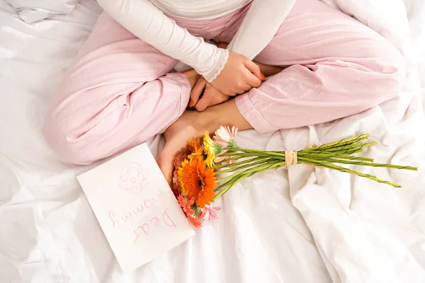Vista recortada de la mujer sentada en la cama cerca del ramo de gerberas y la tarjeta de día de las madres - foto de stock