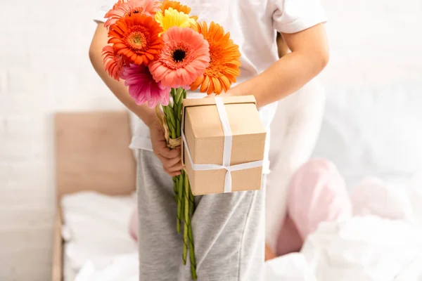 Rückansicht des Jungen mit Blumen und Muttertagsgeschenk in der Nähe der Mutter, die auf dem Bett sitzt — Stockfoto