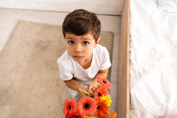 Overhead view of adorable boy looking at camera while holding bouquet of gerberas on mothers day — Stock Photo