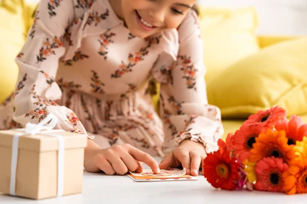 Enfoque selectivo de lindo niño haciendo la tarjeta del día de las madres mientras está sentado cerca de flores y caja de regalo - foto de stock
