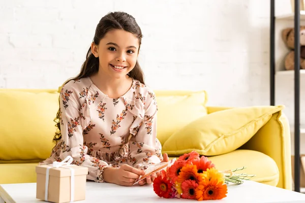 Fröhliches Kind in elegantem Kleid, das in die Kamera blickt, während es am Tisch mit Blumen und Geschenkbox sitzt — Stockfoto