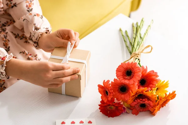 Vista cortada da fita de amarração da criança na caixa de presente do dia das mães enquanto sentado perto de flores na mesa — Fotografia de Stock