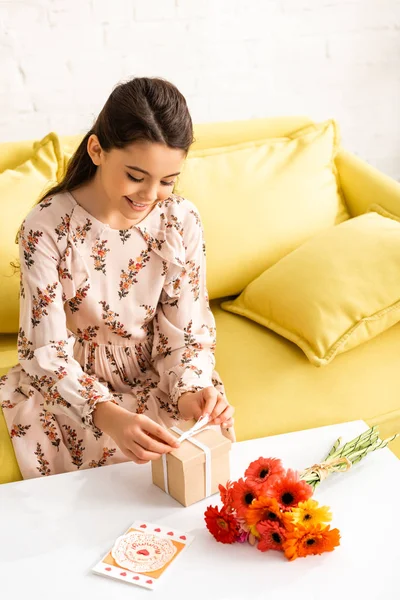 Smiling child in elegant dress tying ribbon on gift box while sitting near flowers and mothers day card — Stock Photo