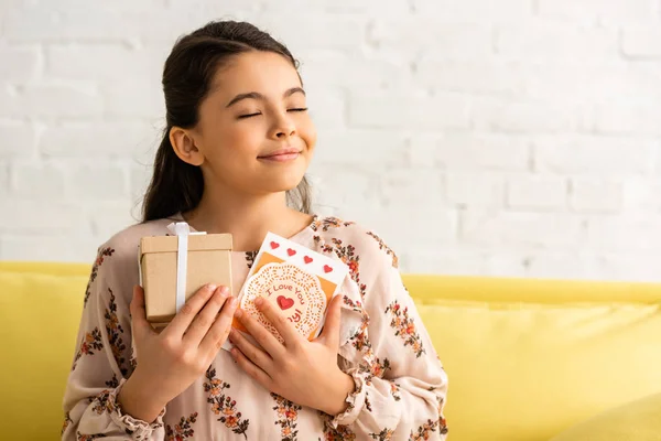 Niño feliz en vestido elegante sentado con los ojos cerrados y la celebración de la caja de regalo y la tarjeta de día de las madres con te amo mamá inscripción - foto de stock