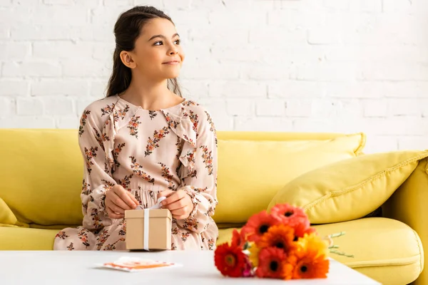 Cute, smiling child in elegant dress sitting near table with flowers and gift box — Stock Photo