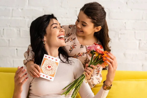 Adorável filha segurando mães cartão de dia e flores enquanto abraçando mãe feliz — Fotografia de Stock