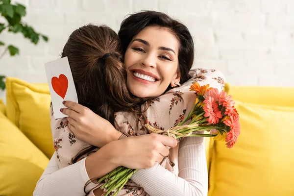 Happy woman holding flowers and mothers day card with heart symbol while hugging adorable daughter — Stock Photo