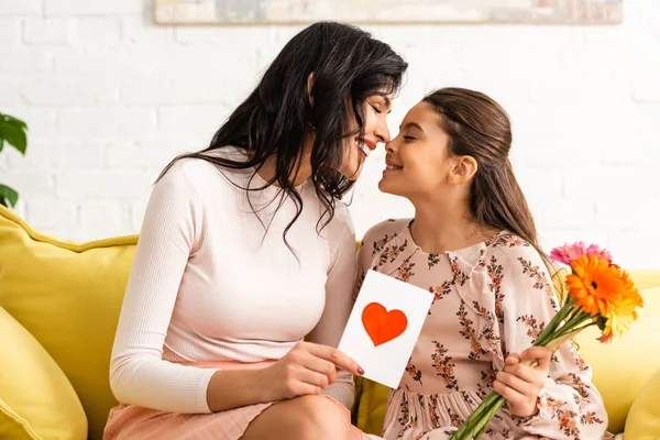 Glückliche Frau mit Muttertagskarte mit Herzsymbol und Tochter mit Blumen auf dem Sofa sitzend und von Angesicht zu Angesicht lächelnd — Stockfoto