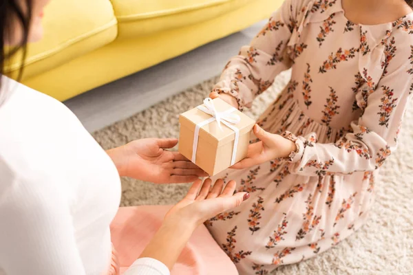 Visão parcial da criança elegante apresentando o presente do dia das mães para a mãe enquanto sentados no chão juntos — Fotografia de Stock